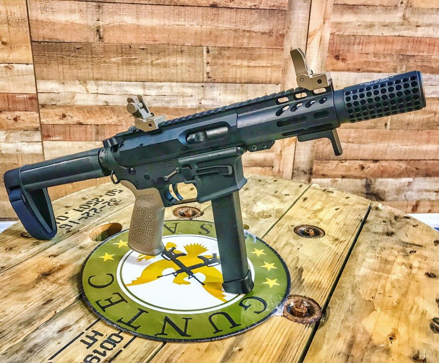 Black pistol-caliber carbine with suppressor on rustic wood, surrounded by ammo; tactical display setting.