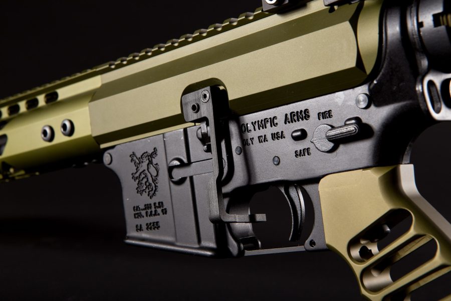 Close-up of tactical AR-style rifle receiver in tan finish with black accents, against dark background.