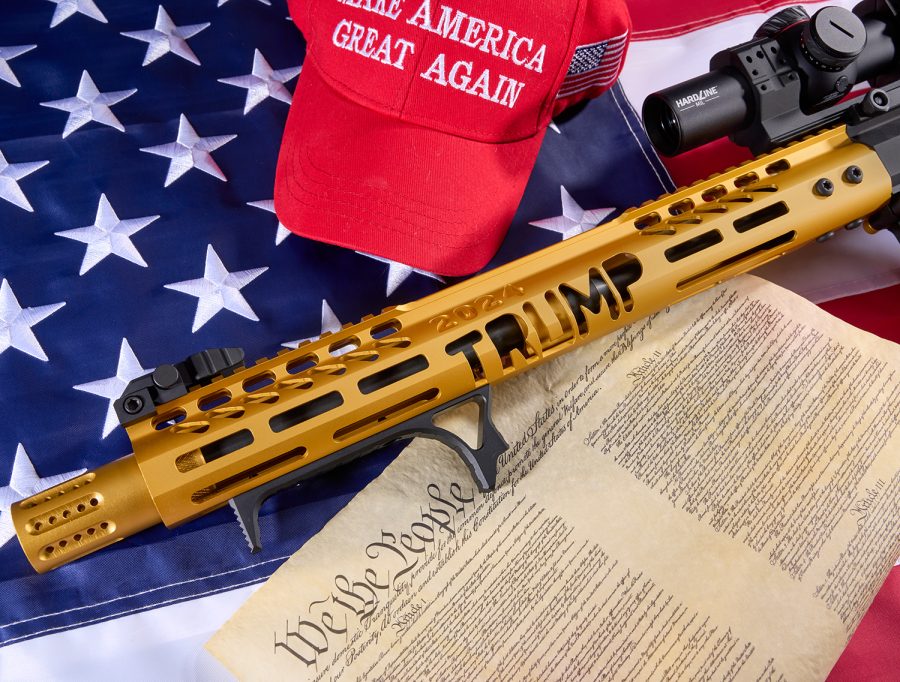 Gold Trump rifle atop American flag, MAGA hat, and U.S. Constitution image.
