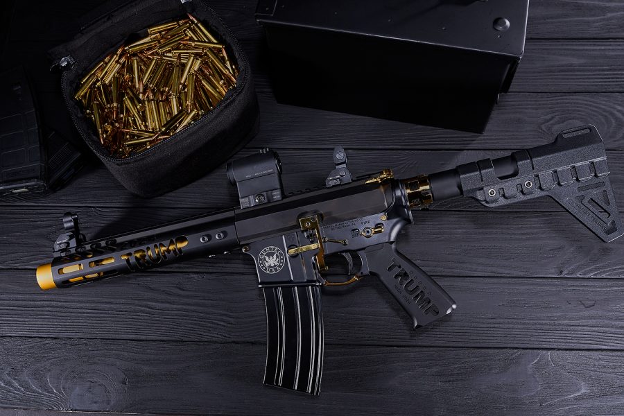 Customized gold-accented Trump AR-15 with optics, ammo, and case on dark surface.