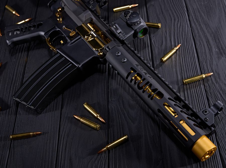 Gold-accented AR rifle with red dot sight on wood, surrounded by brass cartridges.