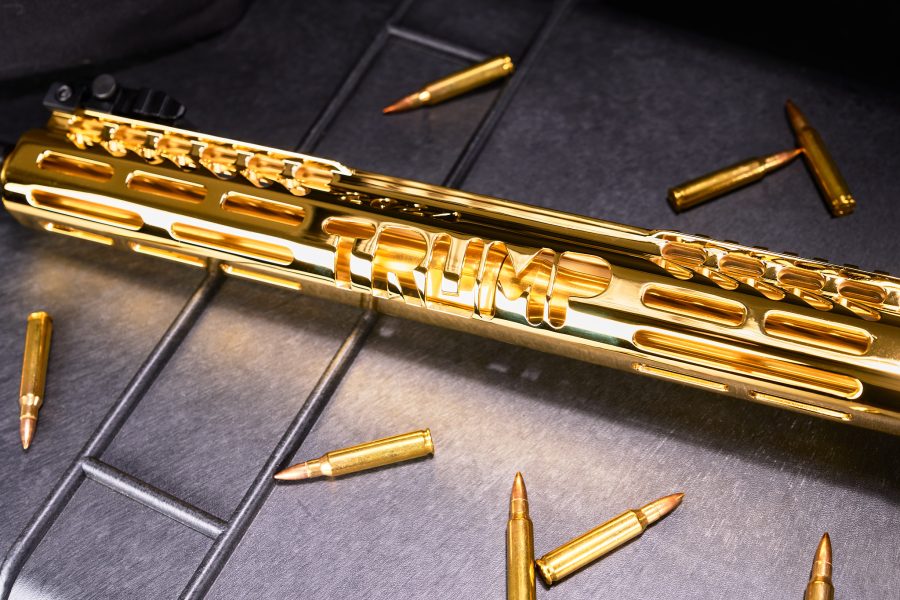 Gold-plated Trump rifle with engraved details, surrounded by matching ammunition on a dark surface.