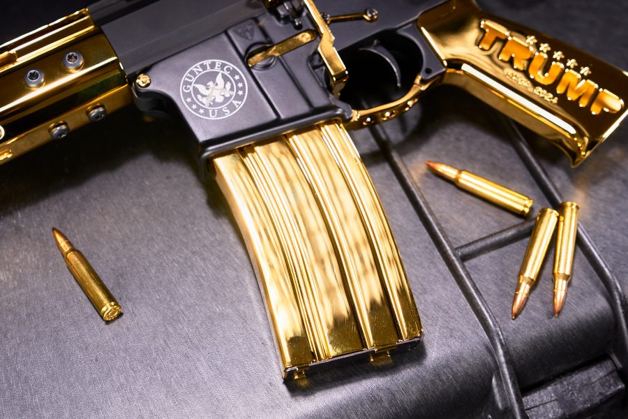 Luxurious gold-plated AR-15 rifle with Trump branding and matching ammunition on dark background.