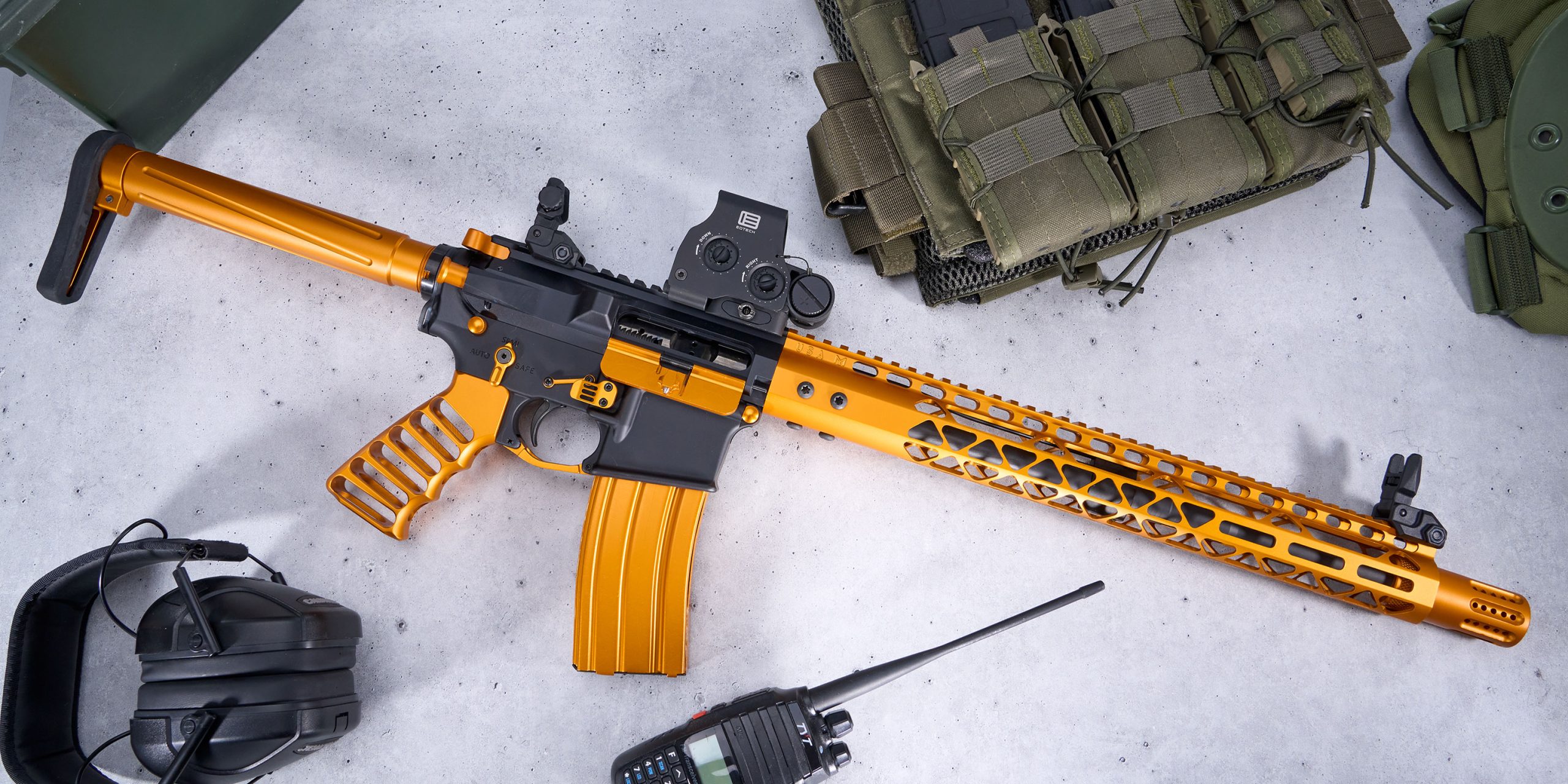 Black and gold AR rifle with tactical gear, including vest, earmuffs, and radio on gray surface.