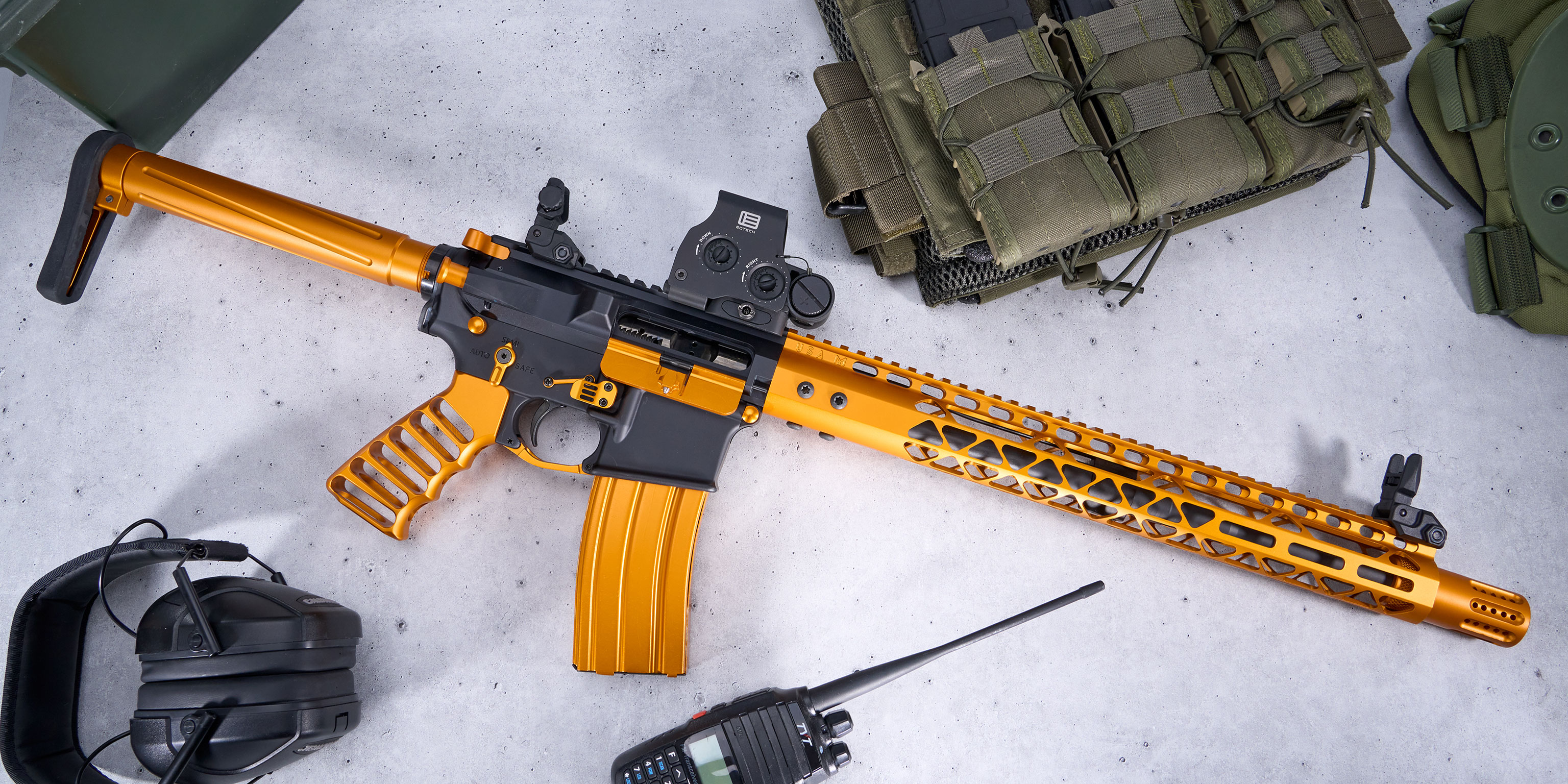Black and gold AR rifle with tactical gear, including vest, earmuffs, and radio on gray surface.