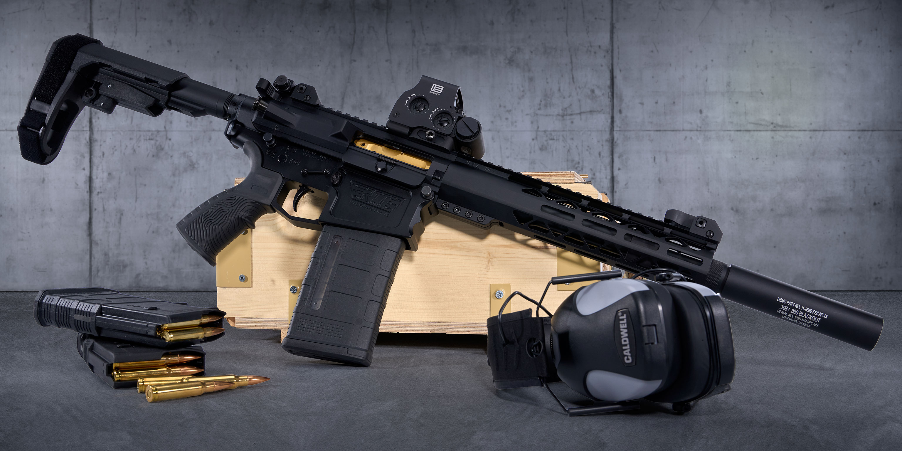 Compact AR-style firearm with accessories and ear protection on a wooden case.